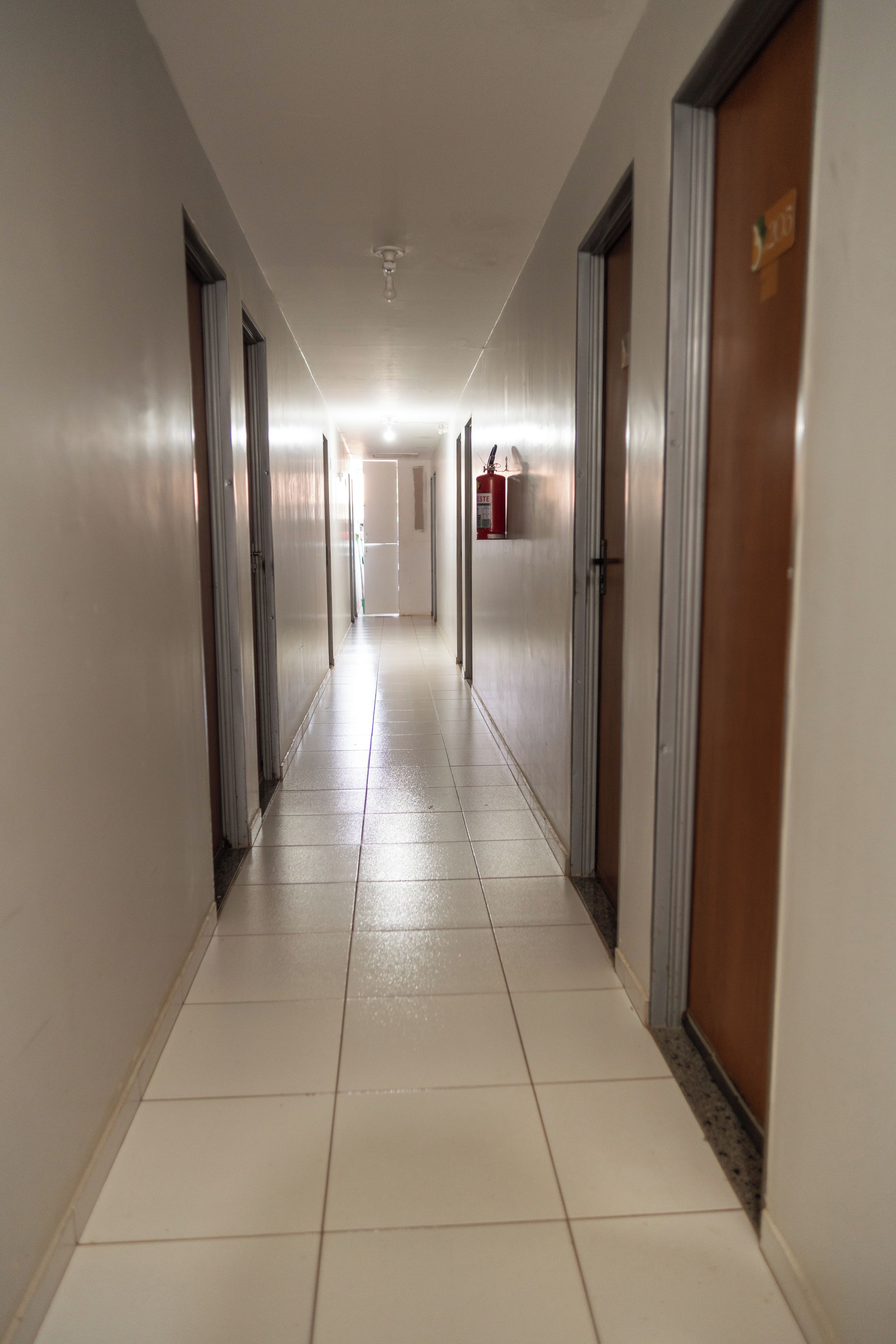 A long, narrow hotel corridor with doors on both sides, a fire extinguisher mounted on the wall, and a ceiling light fixture.