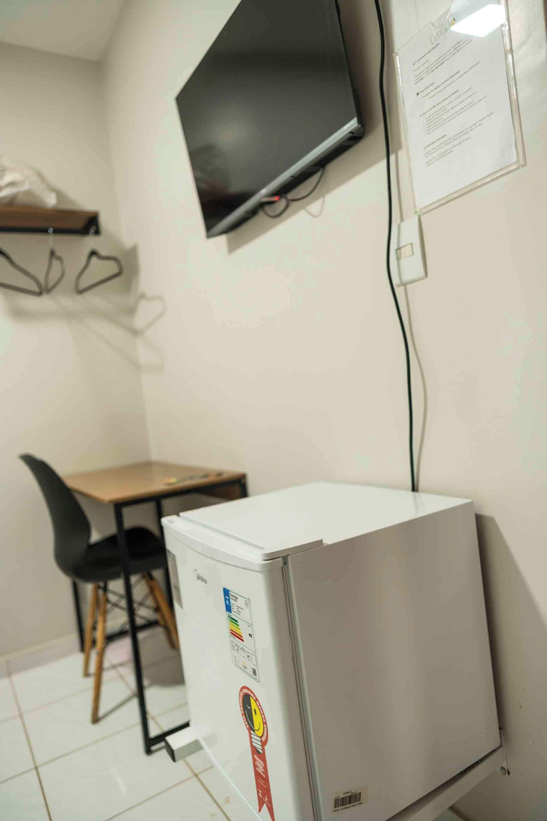 A compact kitchenette with a refrigerator, a small table, and a chair. A flat-screen TV is mounted on the wall above the table.