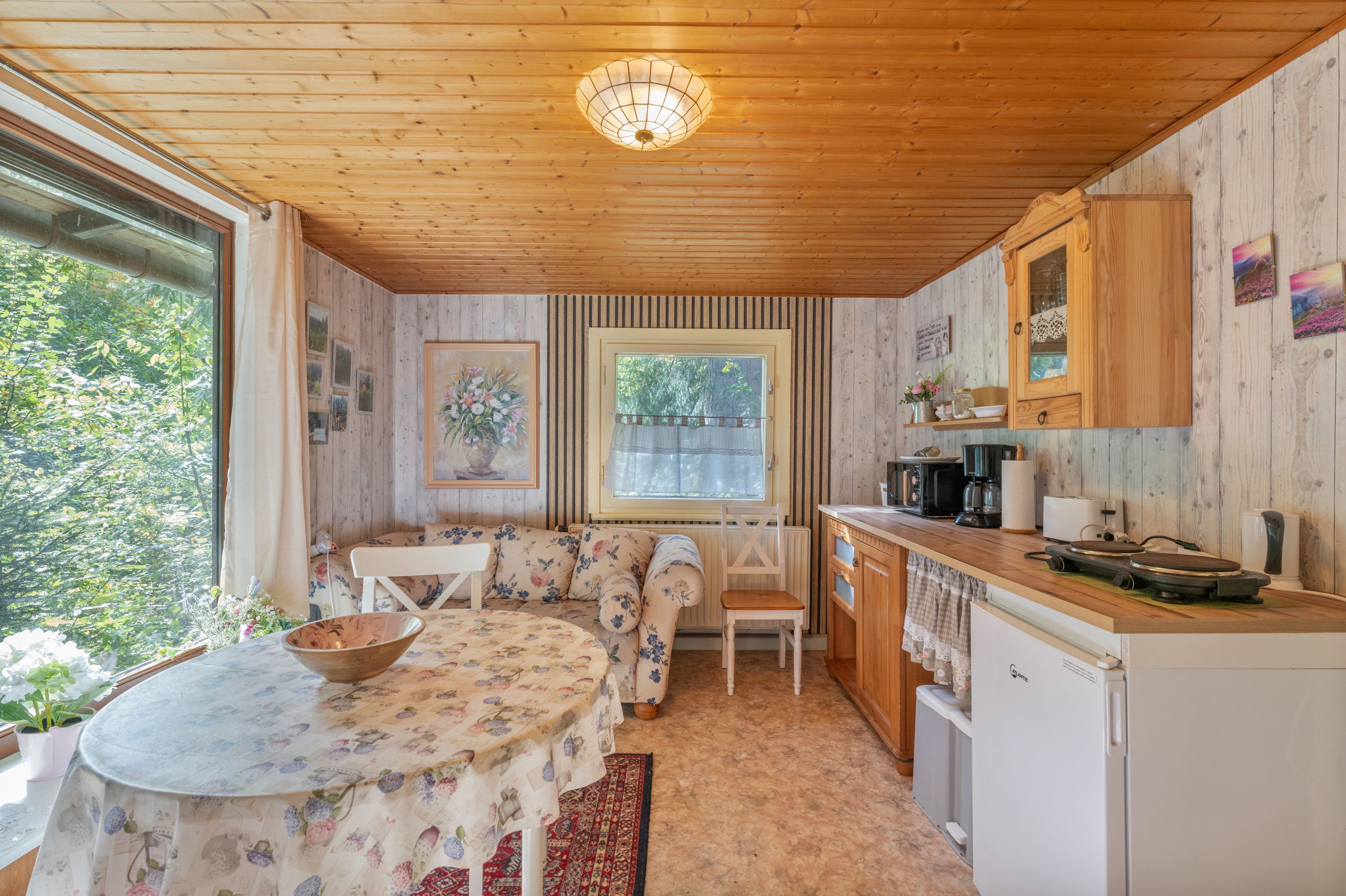 A cozy kitchen with wooden cabinets, a dining table with floral tablecloth, and a window with a view of greenery.