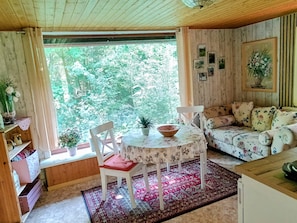Une pièce chaleureuse avec un plafond en bois, un canapé recouvert d’un tissu à motifs floraux, une table ronde avec un saladier, et une fenêtre donnant sur les arbres.