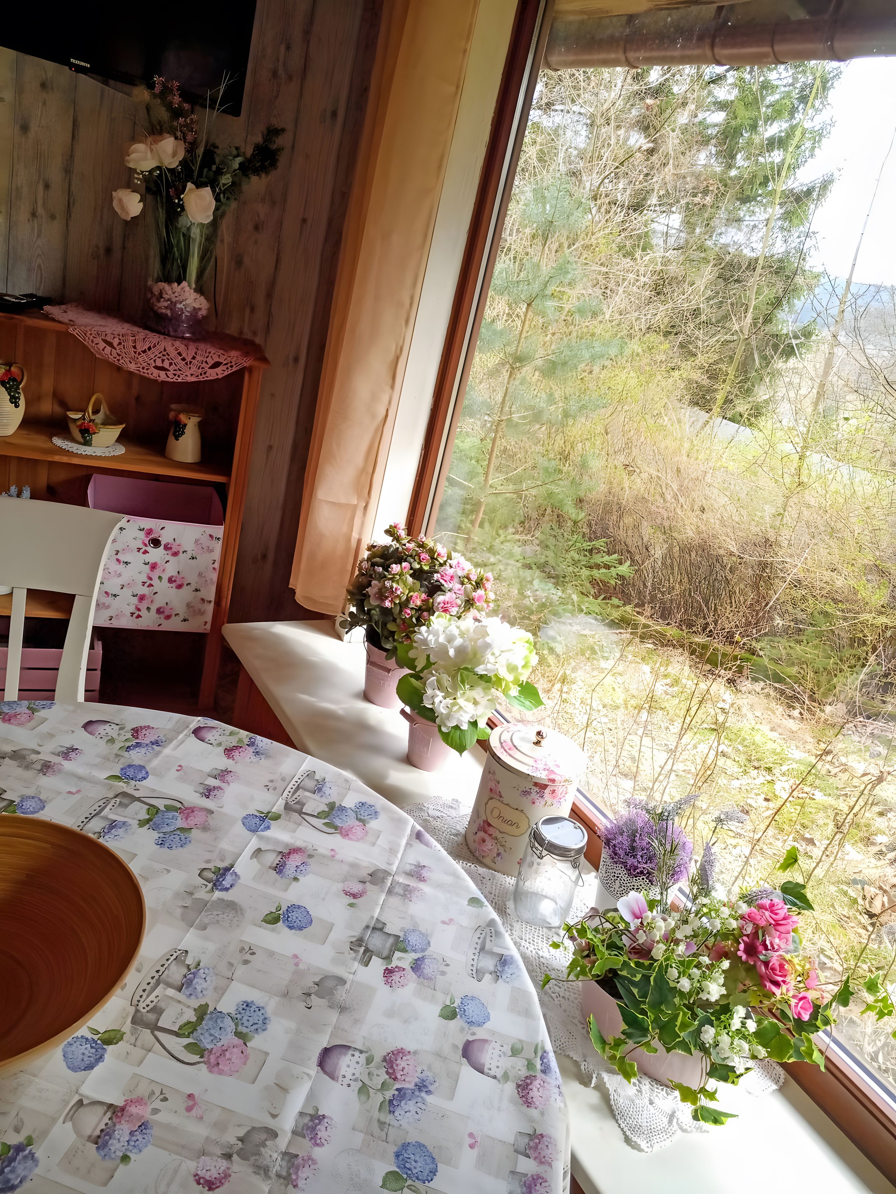 Ein Zimmer mit Holzeinrichtung, einem Tisch mit einer blumigen Tischdecke und verschiedenen Blumentöpfen.