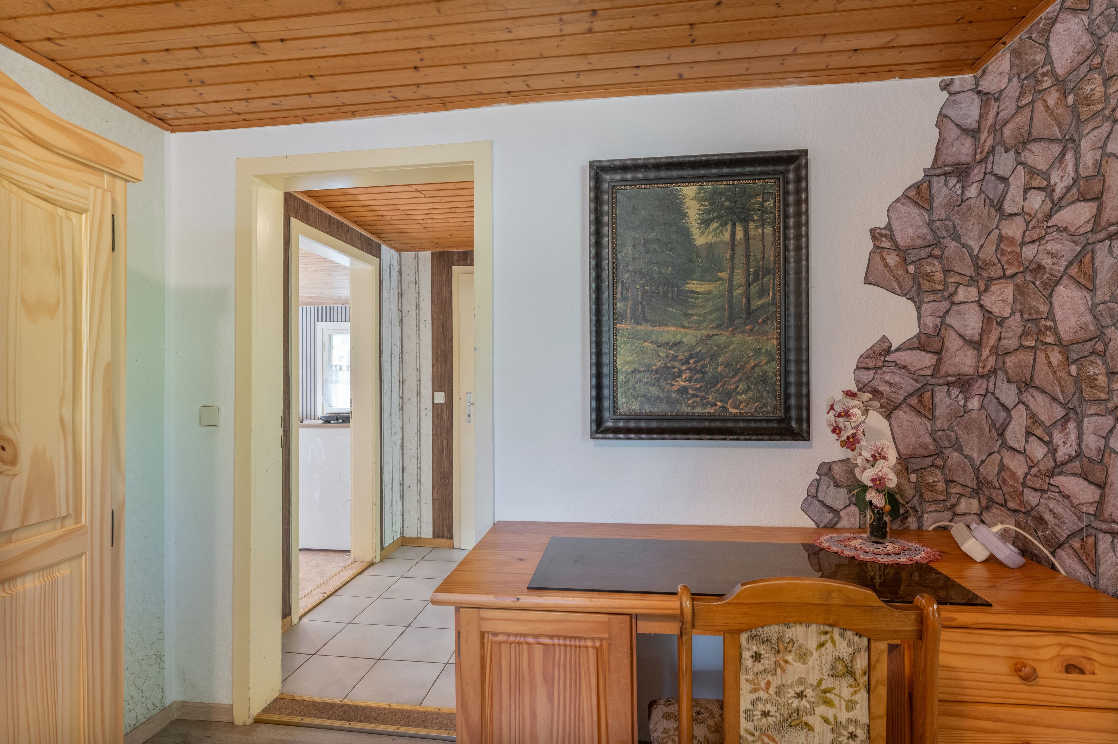 Una habitación con techo de madera, pared de piedra, un cuadro enmarcado, un escritorio con una silla y una puerta que da a otra habitación.