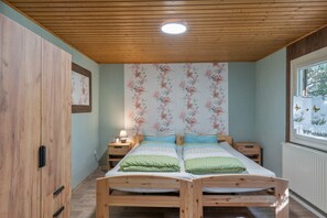 Ein Schlafzimmer mit Holzboden, Blumenmuster-Tapete, zwei Betten mit grüner Bettwäsche, einem Nachttisch mit Lampe und einem Fenster mit Blick auf Bäume.