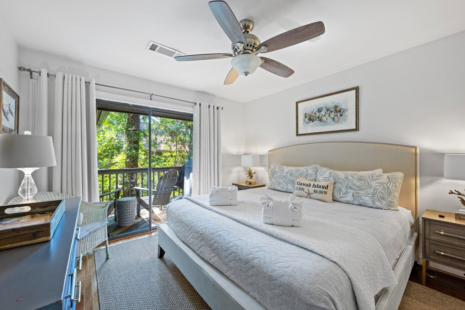 Habitación de hotel con una cama grande, ventilador de techo, balcón con una silla y vista a zonas verdes.