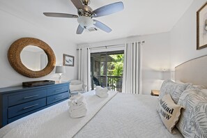 A bedroom with a bed, a ceiling fan, a round mirror, a dresser, and a view of the outdoors. - Kiawah Exclusives | 33 Inlet Cove (Kiawah Island)