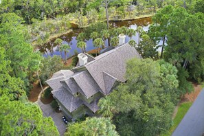 A two-story house with a sloped roof, surrounded by dense greenery and a body of water. - Kiawah Exclusives | 50 Salt Cedar (Kiawah Island)