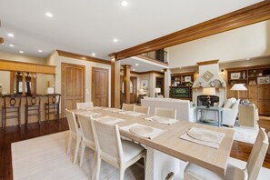 A spacious dining area with a long table and chairs, a kitchen in the background, and a living area with a sofa and television. - Kiawah Exclusives | 50 Salt Cedar (Kiawah Island)
