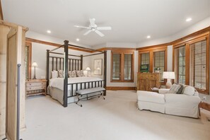 A bedroom with a four-poster bed, a sitting area with a sofa, and large windows with wooden shutters. - Kiawah Exclusives | 50 Salt Cedar (Kiawah Island)