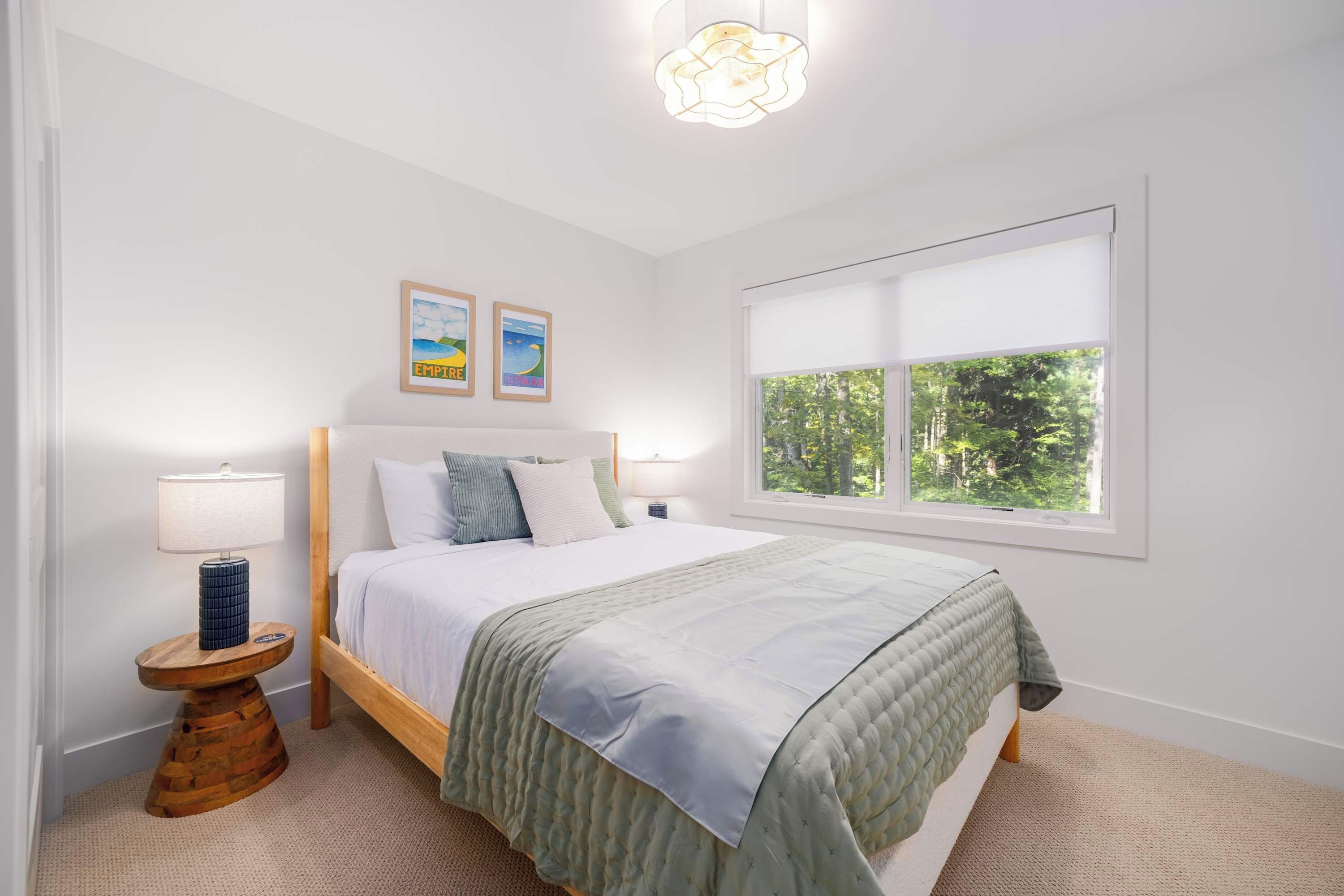 A bedroom with a bed, a nightstand, a lamp, a window with a view of trees, and two framed pictures on the wall.