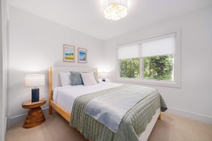 Une chambre à coucher avec un lit, une table de chevet, une lampe, une fenêtre donnant sur des arbres et deux tableaux encadrés accrochés au mur.