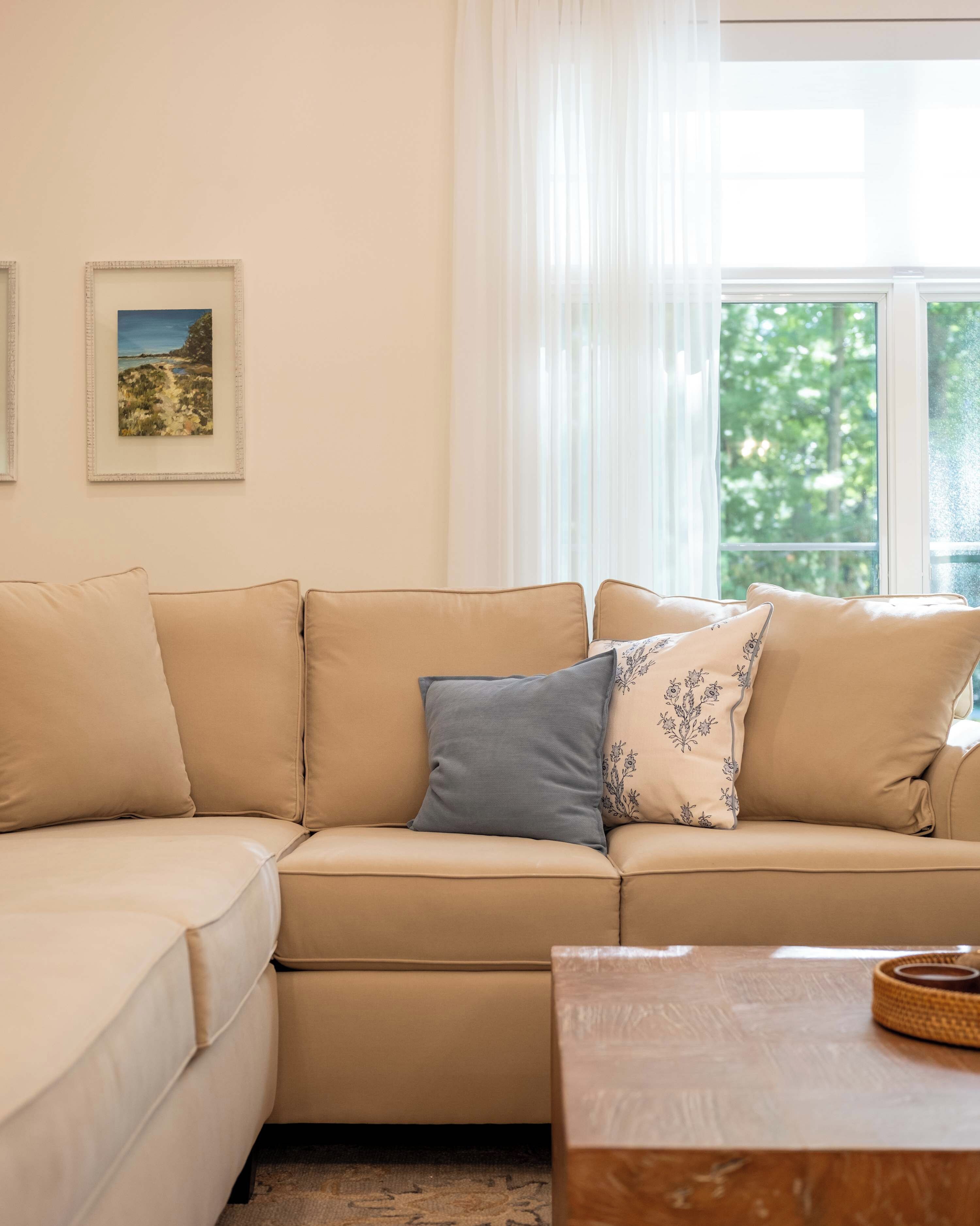 A beige sofa with multiple cushions, a framed picture on the wall, and a window with sheer curtains.