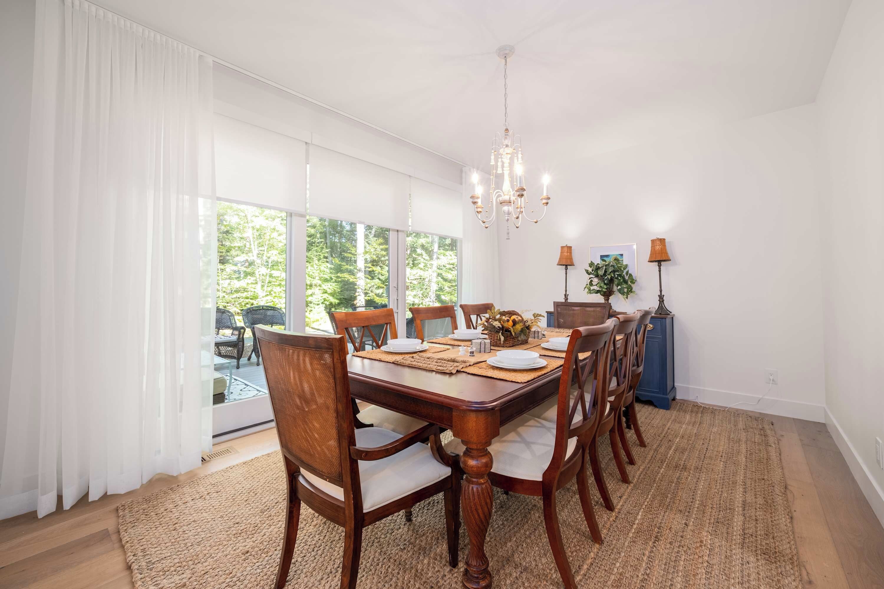 A dining room with a large table, chairs, a chandelier, and a view of greenery outside.