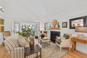 A spacious living room with a fireplace, comfortable seating, and a view through large glass doors. - Kiawah Exclusives | 447 Oceanwoods Cottage (Kiawah Island)