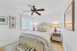 A bedroom with a bed, two bedside lamps, a ceiling fan, a window with blinds, and framed pictures on the wall. - Kiawah Exclusives | 447 Oceanwoods Cottage (Kiawah Island)