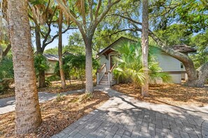 A residential property with a paved driveway, a wooden staircase leading to a house, and surrounding trees. - Kiawah Exclusives | 447 Oceanwoods Cottage (Kiawah Island)