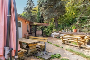 An outdoor patio area with wooden benches and tables, surrounded by greenery and trees.