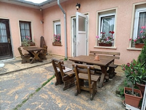 Een terras met een houten tafel en stoelen, een deur en ramen met bloembakken.