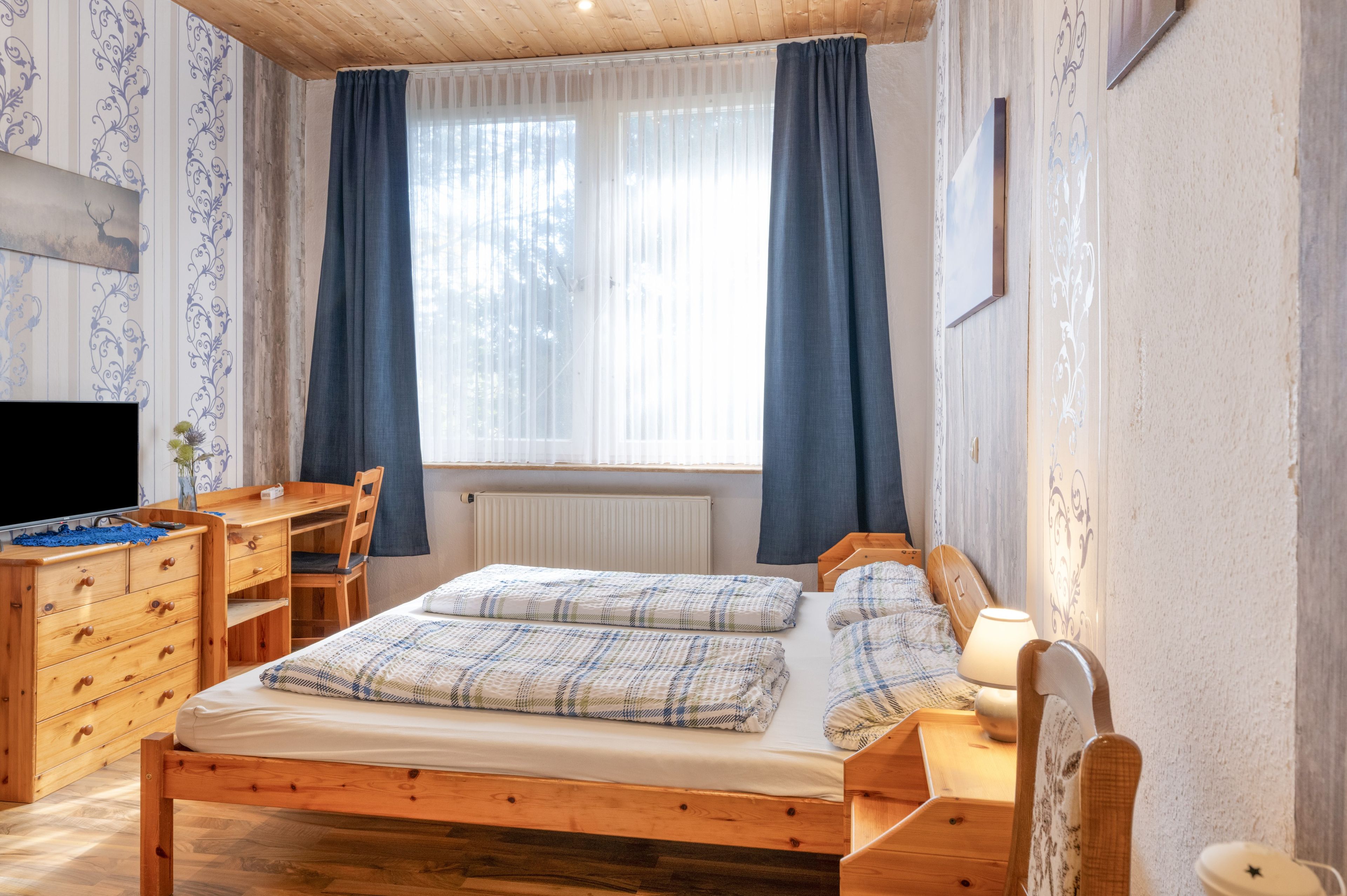 Ein Holzbett mit zwei Kissen, ein Nachttisch mit Lampe, ein Schreibtisch mit Stuhl und ein Fernseher.