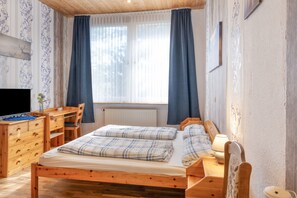 A wooden bed with two pillows, a bedside table with a lamp, a desk with a chair, and a TV.