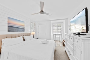 A hotel room with a bed, a television, a ceiling fan, a window with blinds, and a painting on the wall. - Seabrook Exclusives | 1354 Pelican Watch (Seabrook Island)