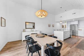 A modern kitchen with white cabinets, a stainless steel refrigerator, and a dining area with a wooden table and black chairs. - Seabrook Exclusives | 1354 Pelican Watch (Seabrook Island)