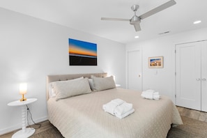 A hotel room with a bed, a ceiling fan, a wall-mounted painting, a bedside table with a lamp, and a door. - Seabrook Exclusives | 1354 Pelican Watch (Seabrook Island)