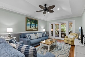 Un salon avec un canapé bleu, une table basse et un fauteuil à rayures. On y trouve également un ventilateur de plafond et un tableau représentant une scène de plage.