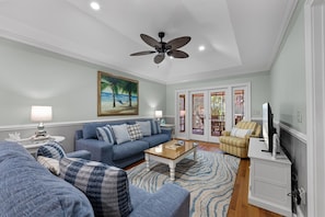A living room with a blue sofa, a coffee table, a striped armchair, a ceiling fan, and a painting of a beach scene. - Seabrook Exclusives | 1151 Summerwind (Seabrook Island)