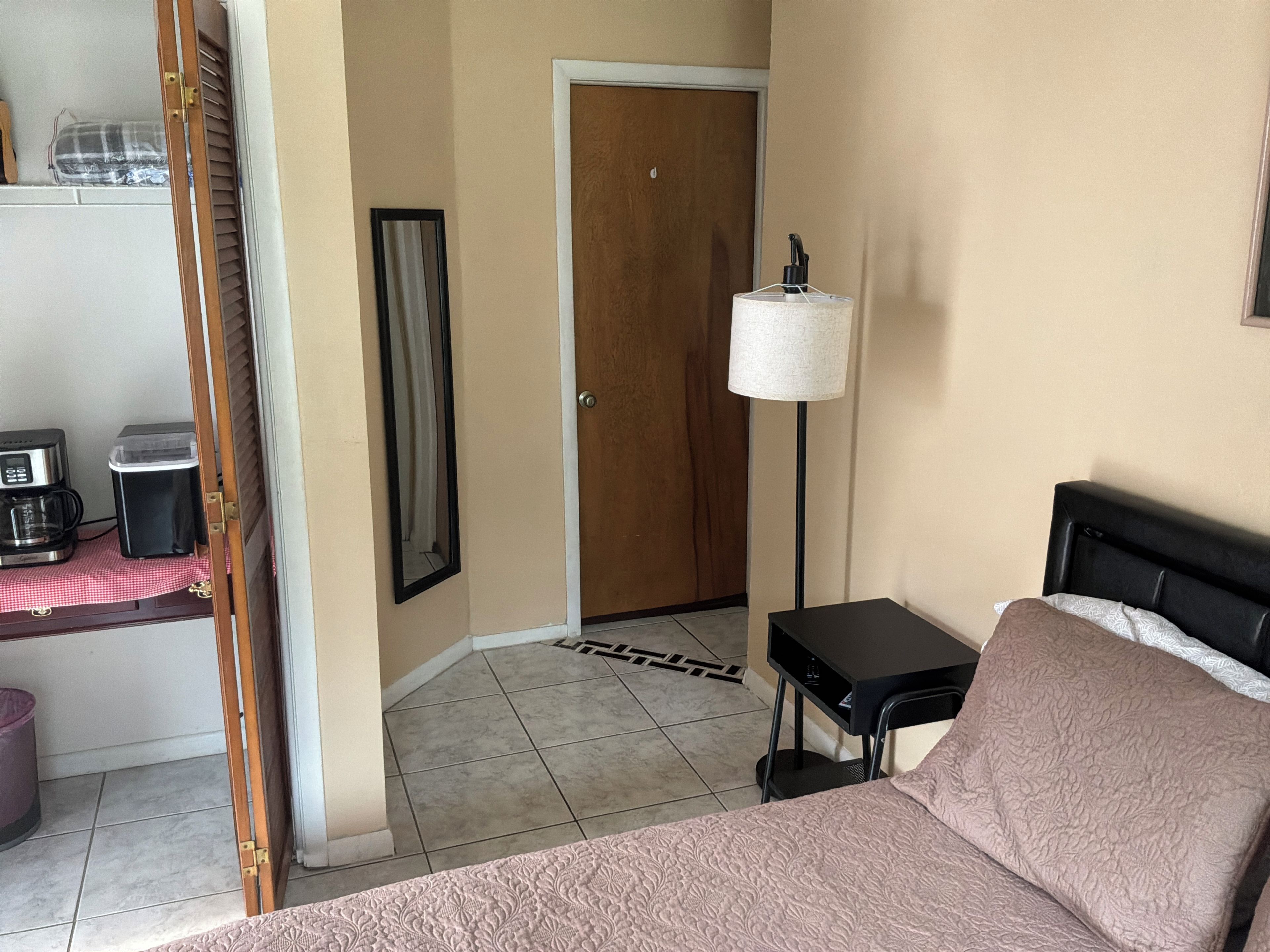 Habitación de hotel con cama, escritorio, cafetera, cesto de basura, puerta y lámpara de pie.