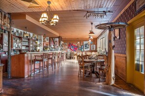 Uma área de bar com piso de madeira, diversos arranjos de assento e variedade de garrafas e copos nas prateleiras.
