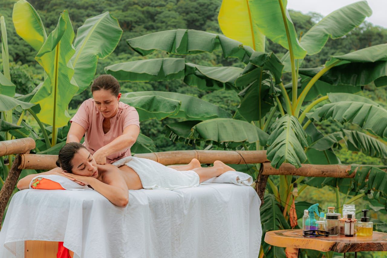 A person receiving a massage on a wooden platform outdoors, surrounded by large banana leaves and greenery.