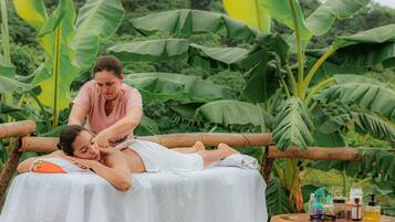 A person receiving a massage on a wooden platform outdoors, surrounded by large banana leaves and greenery.