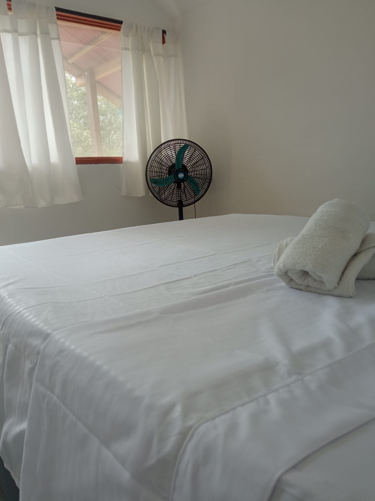 Una cama con ropa blanca, una toalla blanca y un ventilador de pie en una habitación con paredes blancas y una ventana con cortinas translúcidas.
