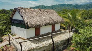 A small, single-story building with a thatched roof, surrounded by lush greenery and a stone pathway leading to it.