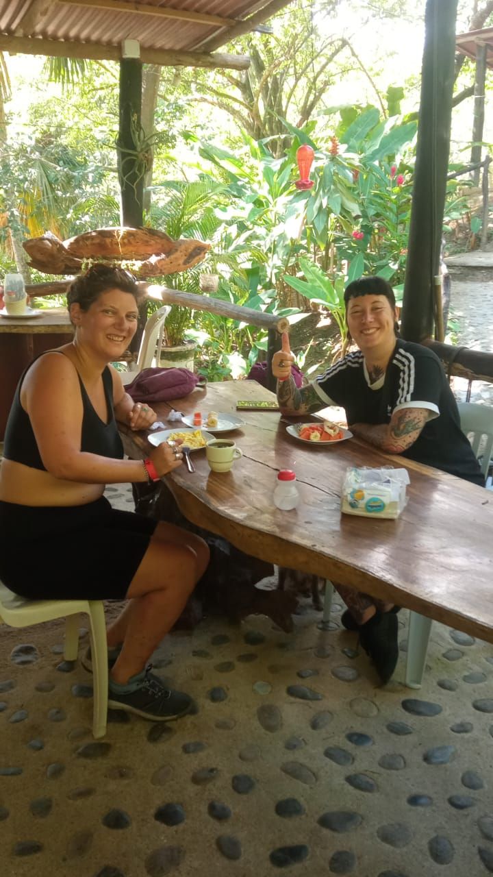 Twee personen zitten aan een houten tafel met eten en drinken, omringd door groen.