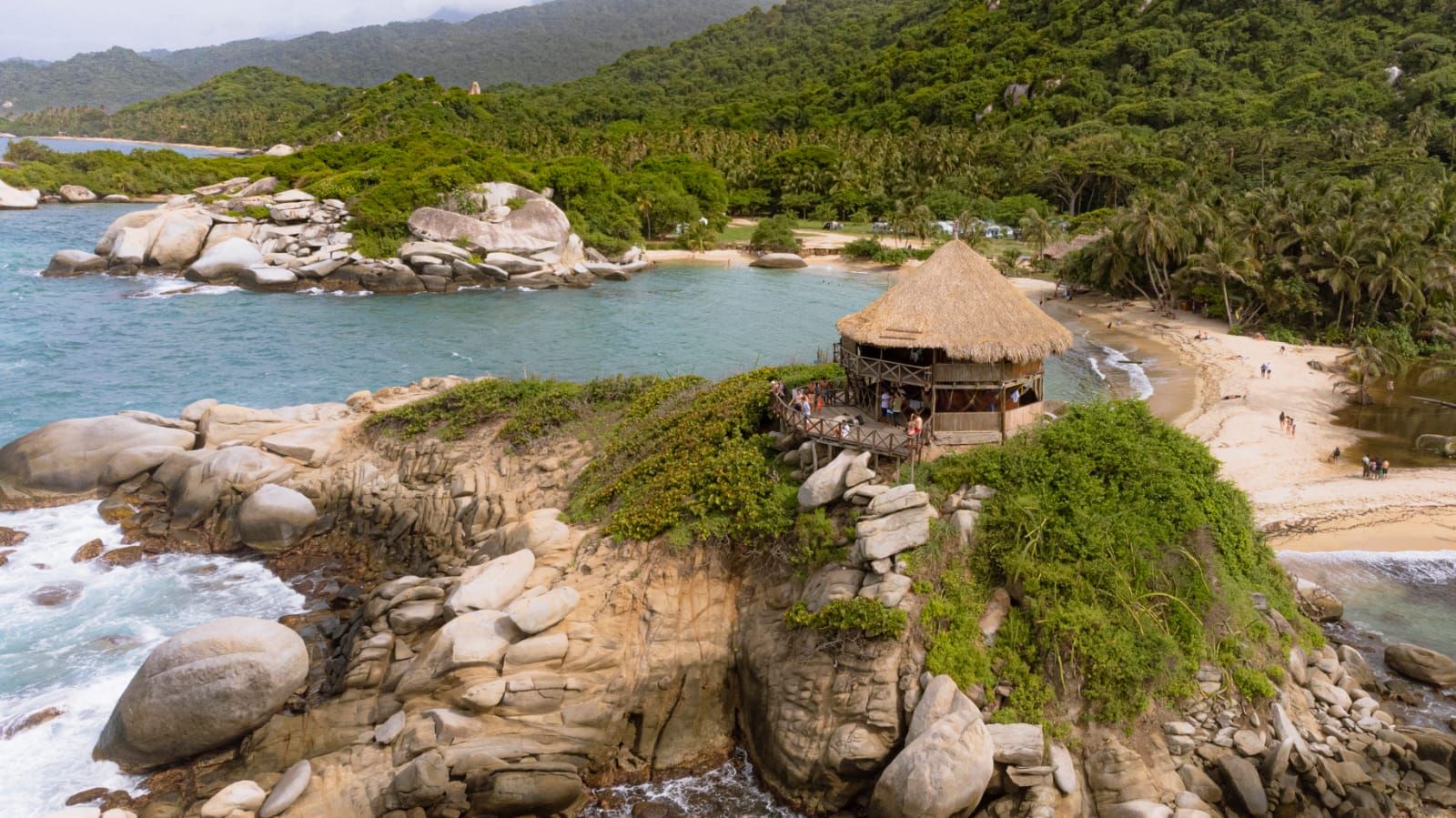 Un paisaje costero con una estructura de techo de paja en un promontorio rocoso, con vista a una playa y al mar.