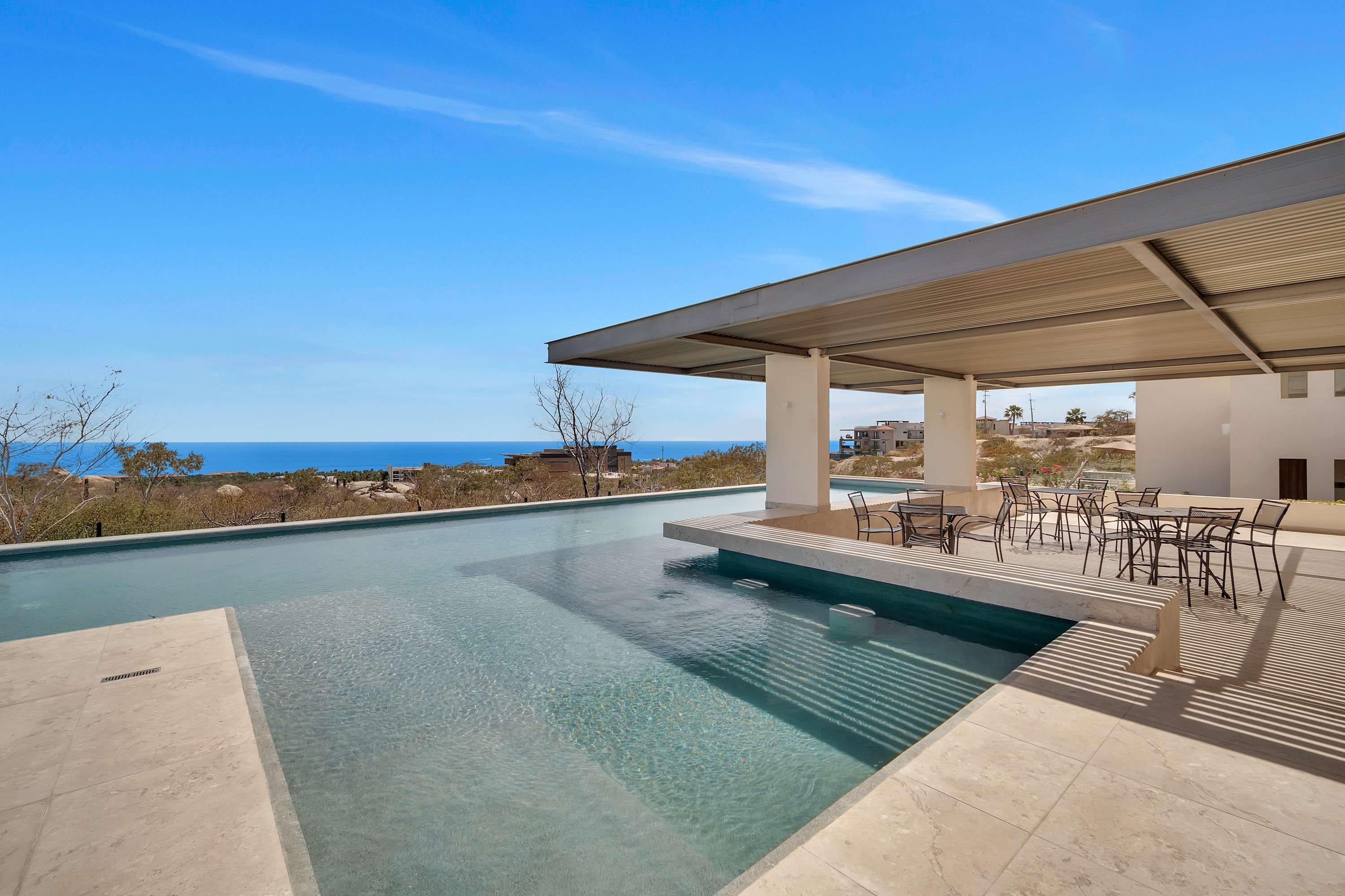Ein moderner Poolbereich im Freien mit klarem, blauem Wasser, einer überdachten Sitzgelegenheit mit Stühlen und Blick auf den Ozean.