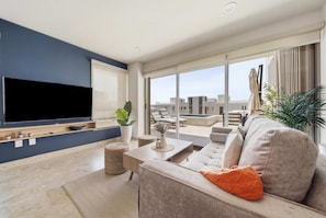 A modern living room with a flat-screen TV, a sofa, a coffee table, and a large window offering a view of the cityscape. - Brand New Pet-friendly Condo - Private Pool (Cabo San Lucas)