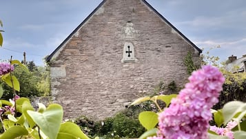 A stone building with a cross on top, surrounded by greenery and flowers.