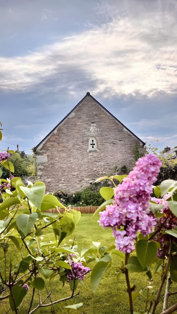 Edificio de piedra con cruz en la parte superior, rodeado de follaje y flores.
