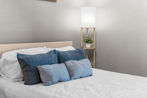 A bed with white bedding and multiple blue pillows, a bedside table with a plant, and a lamp. - Fall Creek Condo | Minutes to Strip, SDC & Lakes (Branson)