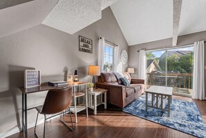 A modern living room with a brown leather sofa, a wooden coffee table, a desk with a chair, and a large window offering a view of the outdoors. - Fall Creek Condo | Minutes to Strip, SDC & Lakes (Branson)