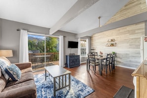 A spacious living room with a large sliding glass door leading to an outdoor area, a flat-screen TV mounted on the wall, and a dining area with a wooden table and chairs. - Fall Creek Condo | Minutes to Strip, SDC & Lakes (Branson)