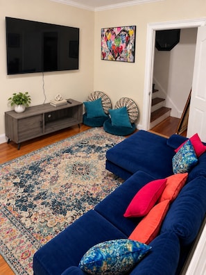 Una sala de estar con un sofá azul, un televisor de pantalla plana, un cuadro abstracto colorido, un tapete estampado y una escalera al fondo.