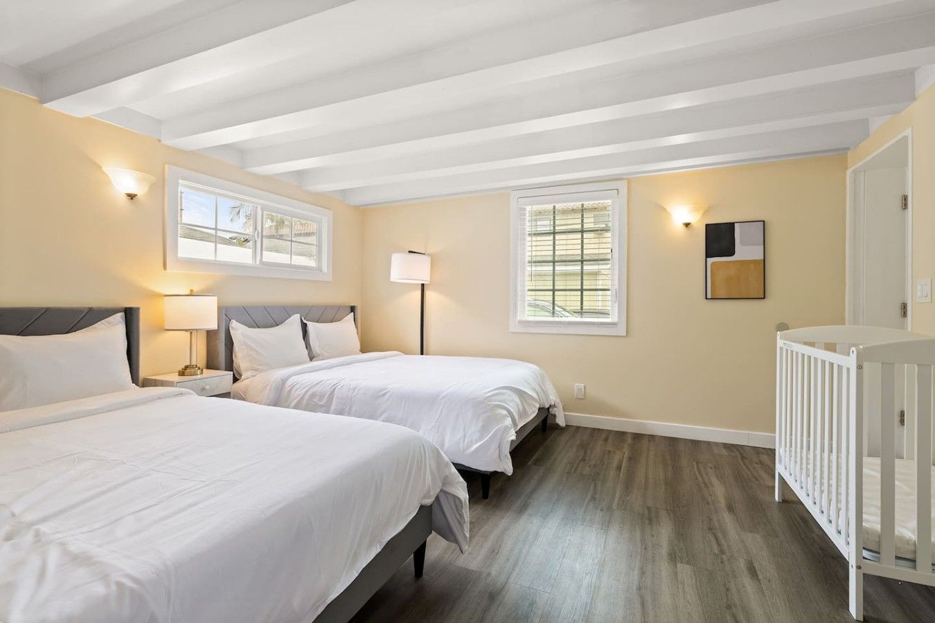 A hotel room with two beds, a crib, a window with blinds, and a wall-mounted lamp.