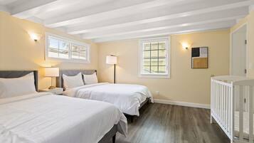 A hotel room with two beds, a crib, a window with blinds, and a wall-mounted lamp.