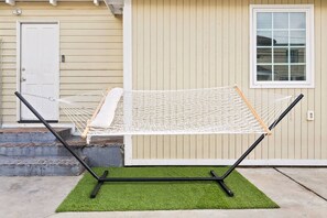 Een hangmat op een betonnen terras, met een groen, synthetisch gazon, een beige huis met een witte deur en raam, en een zwart metalen frame.