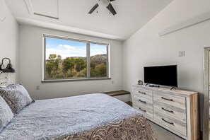 A bedroom with a large window, a bed with patterned bedding, a TV on a wooden dresser, and a ceiling fan. - Quaint Riverfront Property Close to the Resort! Pet Friendly. (Steamboat Springs)