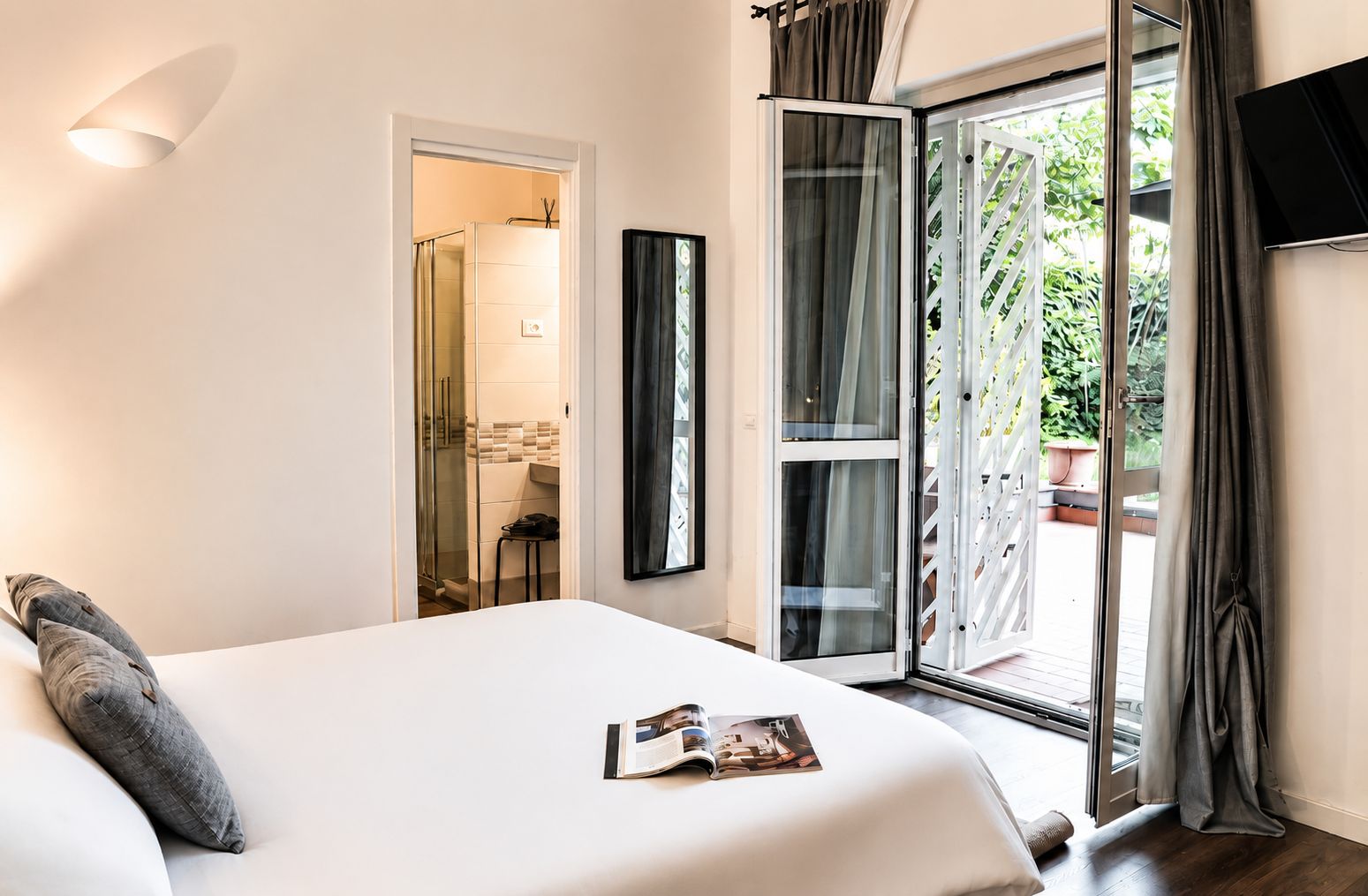 Une chambre d’hôtel avec un lit, un magazine posé sur le lit, une douche, un balcon avec des rideaux et un téléviseur fixé au mur.