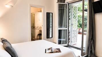 Ein Hotelzimmer mit einem Bett, einer Zeitschrift auf dem Bett, einer Dusche, einem Balkon mit Vorhängen und einem Fernseher an der Wand.